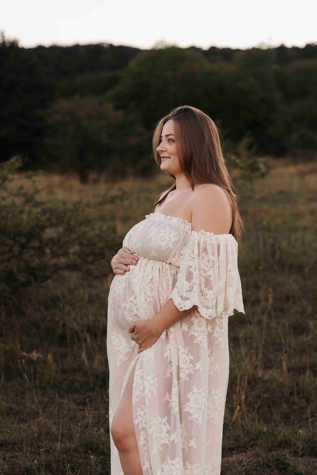 Babybauch Outdoor Fotografie Wien – Lucia im weißen Spitzenkleid, lächelnd mit Hand am Bauch